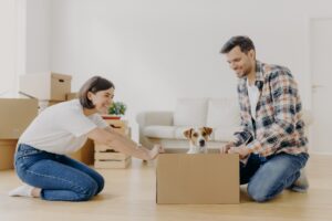 Horizontal shot of lovely woman moves cardboard box with small puppy to husband side